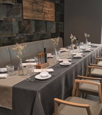 Restorāna galds ar tumši pelēku galdautu, smilškrāsas galda celiņu un baltām salvetēm.
