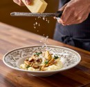Šefpavārs rīvē sieru pār sēņu risoto uz stilīga restorāna porcelāna ar dabīgām nokrāsām.