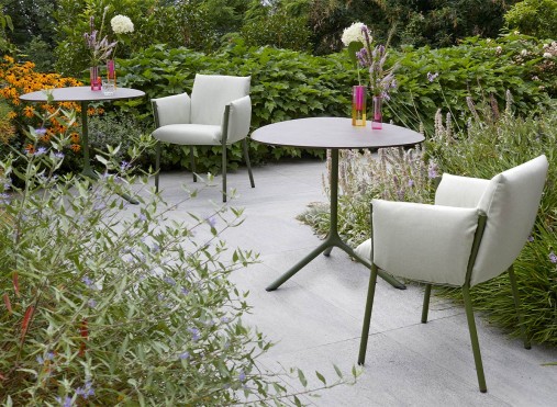 A lush outdoor terrace with light green chairs and tables, surrounded by vibrant greenery and flowers, creating a serene dining atmosphere.